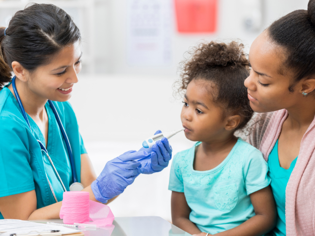 A nurse in teal scrubs takes the temperature of a young girl while the girl sits in her mother's lap.