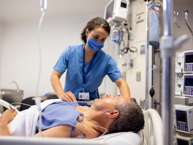 An ICU nurse in blue scrubs who's wearing a face mask treats a male patient.