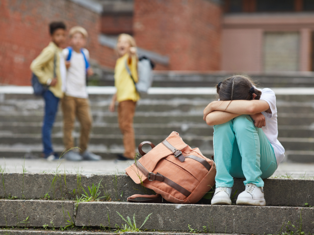 A young child sits on steps outside of a school, lying her head in her hands and her backpack next to her. In the background, a group of children are talking. 
