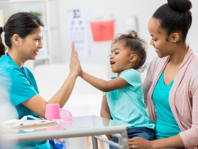 A nurse high-fives a young girl, who's sitting in her mother's lap, after providing pediatric wound care.