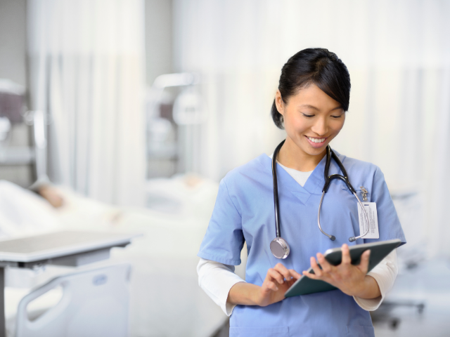 A nurse in a hospital room reviews information on a tablet.