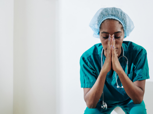 A nurse wearing surgical scrubs sits with her hands resting on the bridge of her nose, representing nurse understaffing.