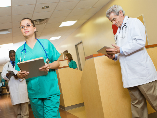A nurse walks through a hospital intake area as a doctor reviews files in the background.